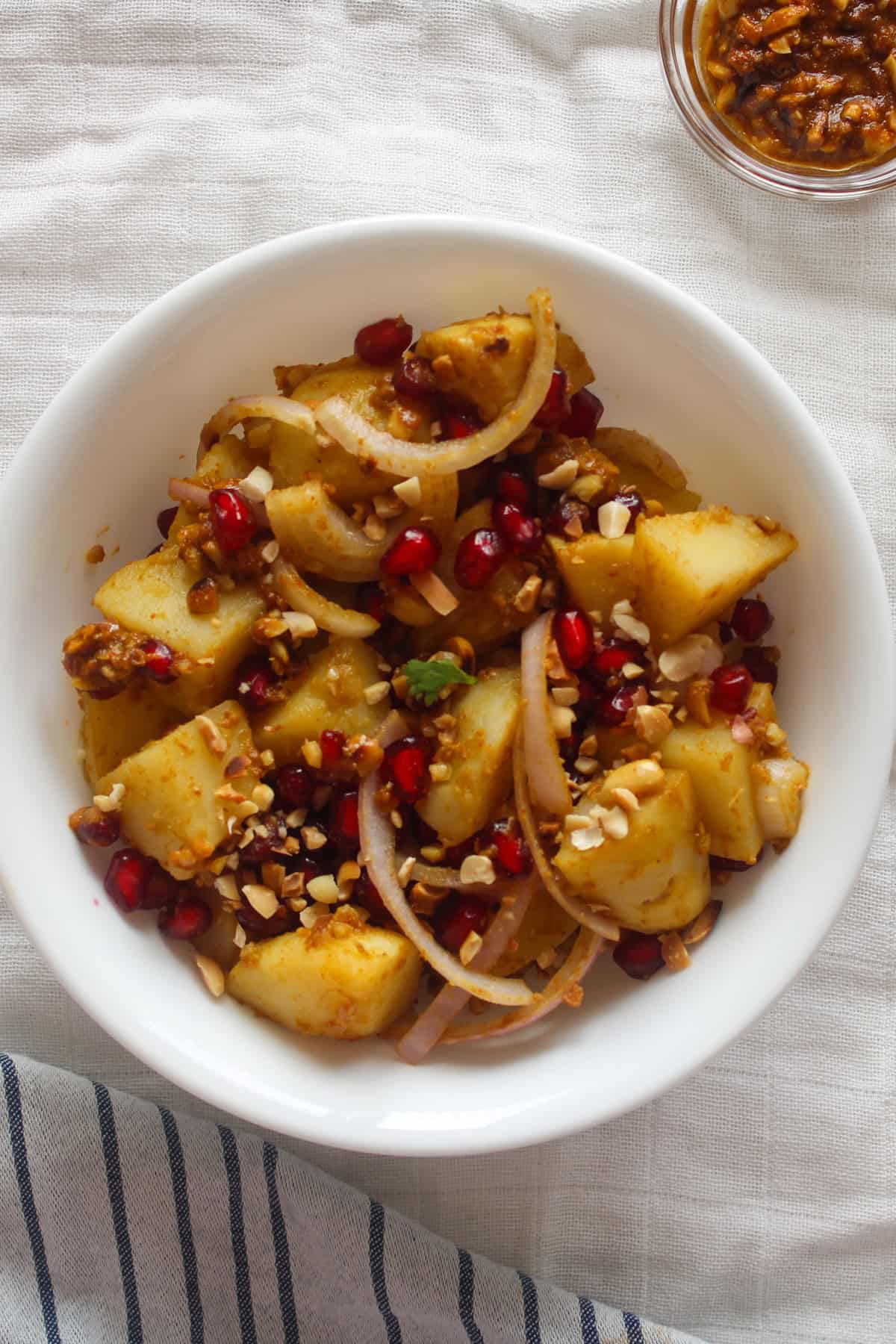 Close overhead view of Indian potato salad with coated potato cubes, red pomegranate seeds, onion slices, and roasted peanuts.