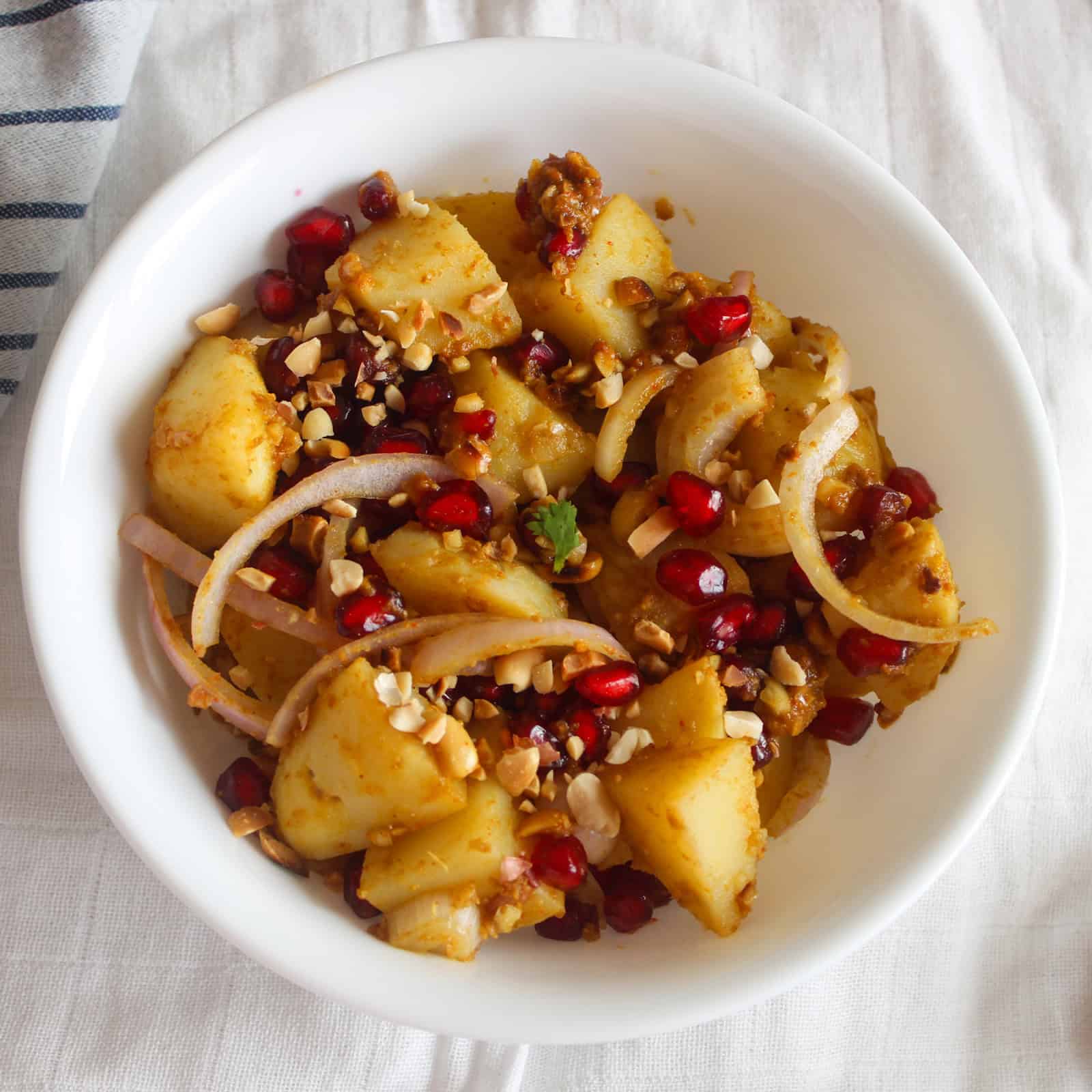 Finished Indian potato salad served with golden potato cubes, bright pomegranate seeds, onion slices, and roasted peanuts.
