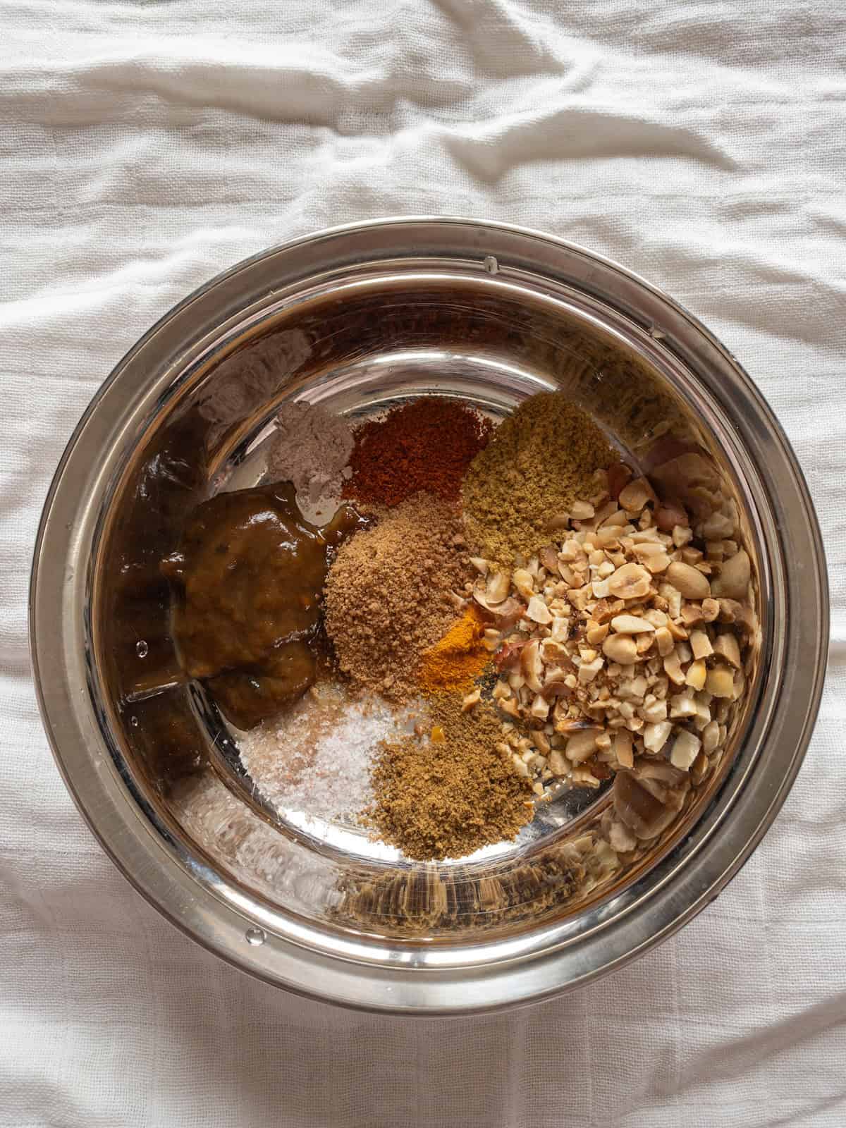 Tamarind paste, jaggery, crushed roasted peanuts, ground spices, and salts arranged in a stainless steel mixing bowl.