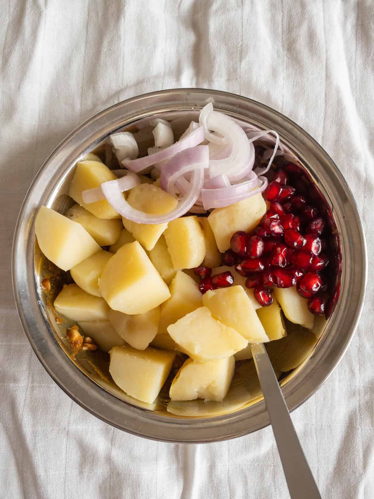 Boiled potato cubes, sliced onion, and pomegranate seeds added over the prepared dressing in a steel bowl.