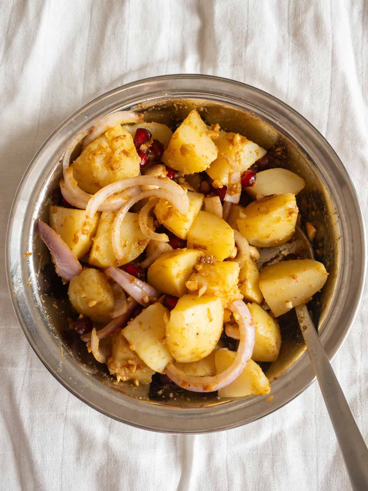 Potato cubes, onion slices, and pomegranate seeds gently tossed with tamarind peanut dressing in a steel bowl.