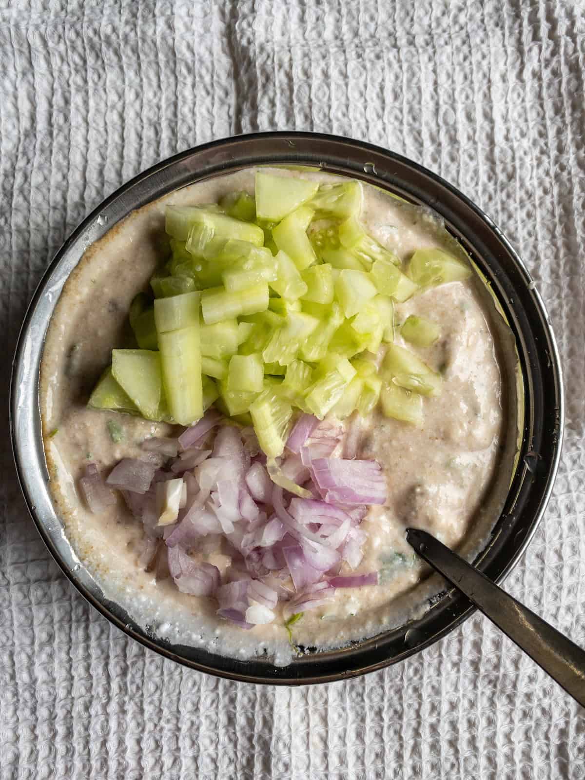 Chopped cucumber and red onion added over the creamy avocado yogurt mixture in a steel bowl.