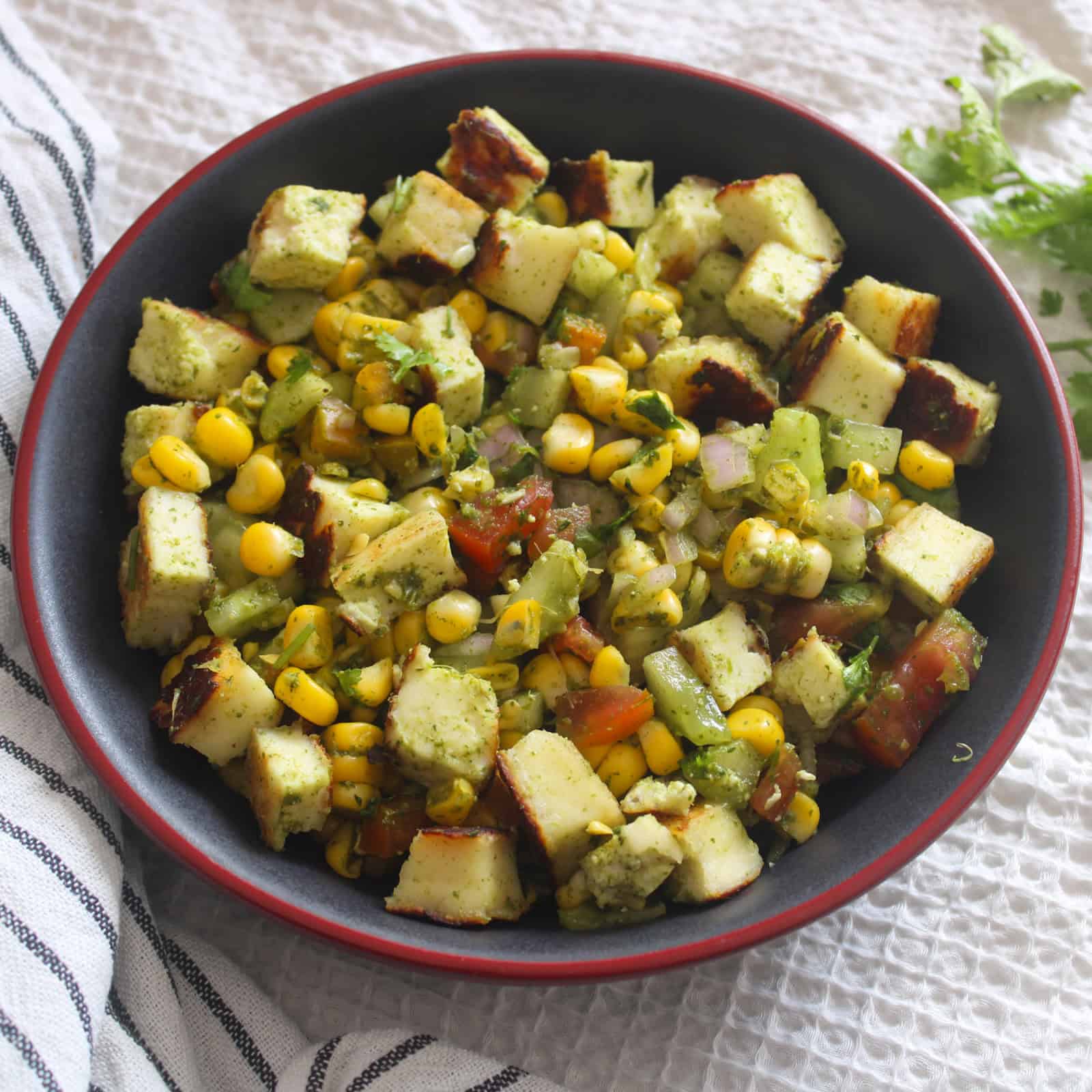 Close view of corn paneer chaat salad with browned paneer, yellow corn, chopped vegetables, and green chutney coating.