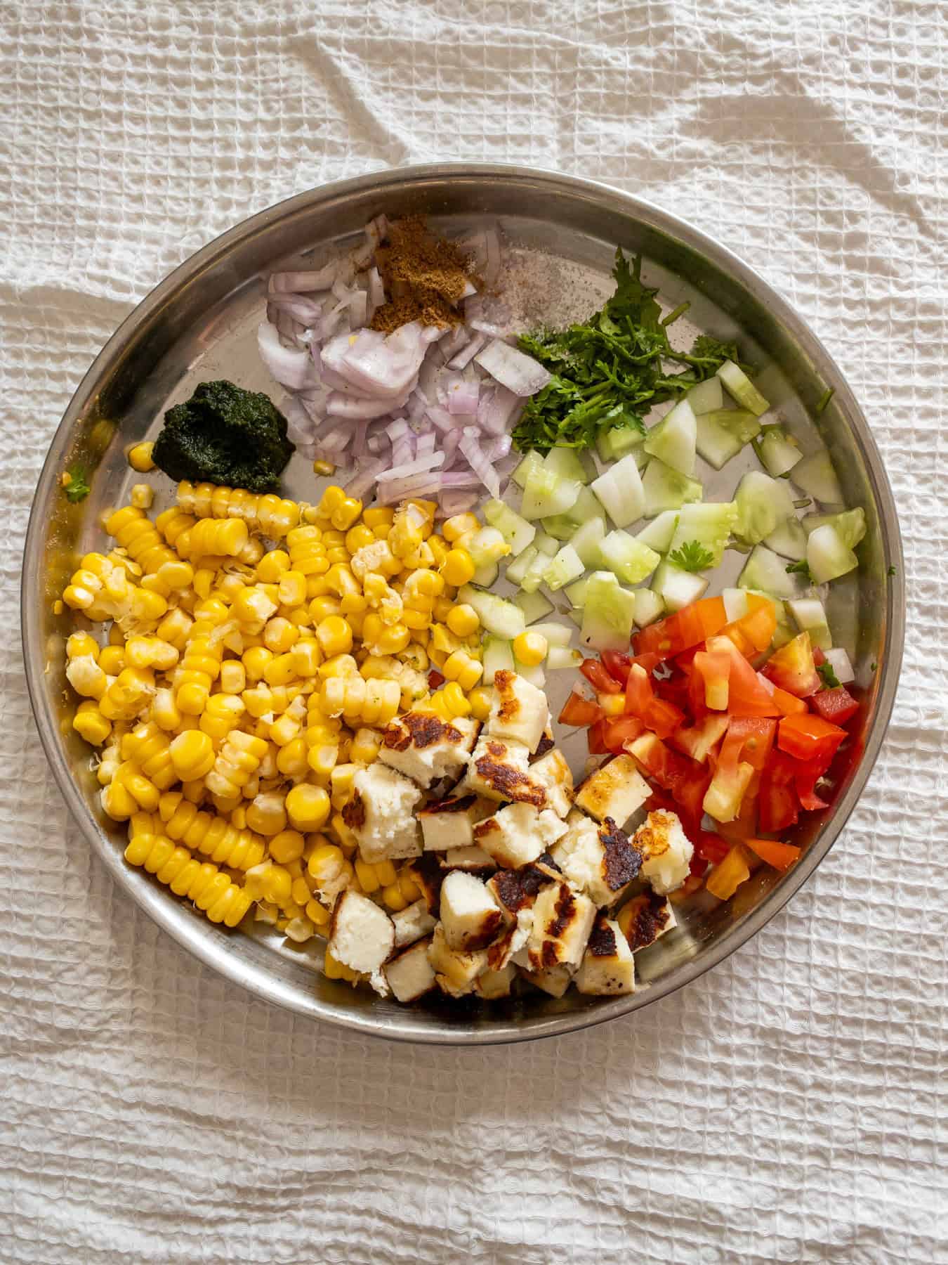 Steamed corn, pan-seared paneer, chopped onion, tomato, cucumber, coriander, green chutney, and seasonings arranged on a steel plate.