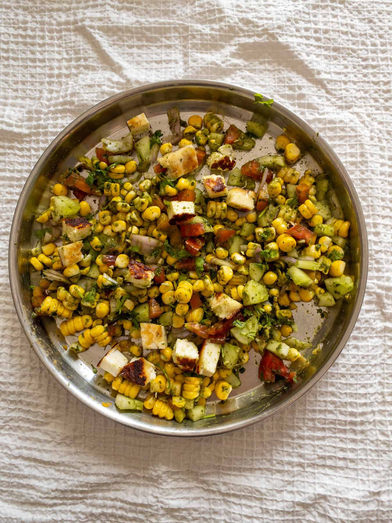 Corn paneer chaat salad tossed with green chutney, vegetables, coriander, and browned paneer cubes on a steel plate.