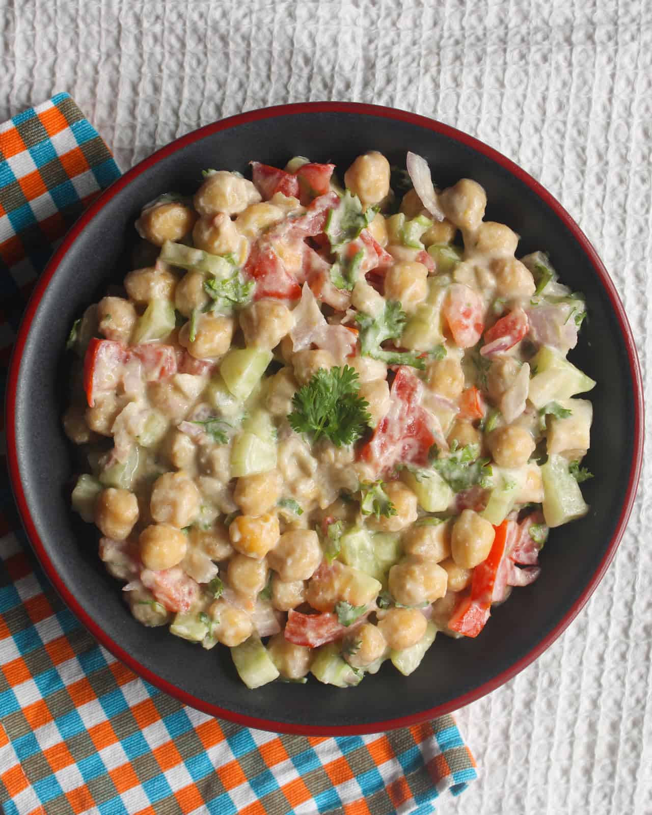 Creamy chickpea salad topped with fresh coriander in a black bowl beside a colorful checked cloth.