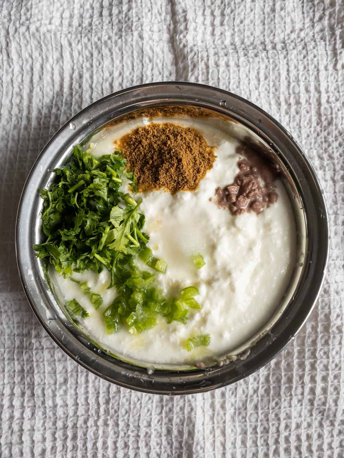 Thick yogurt with mashed avocado, cumin powder, chopped green chili, and coriander in a steel bowl.