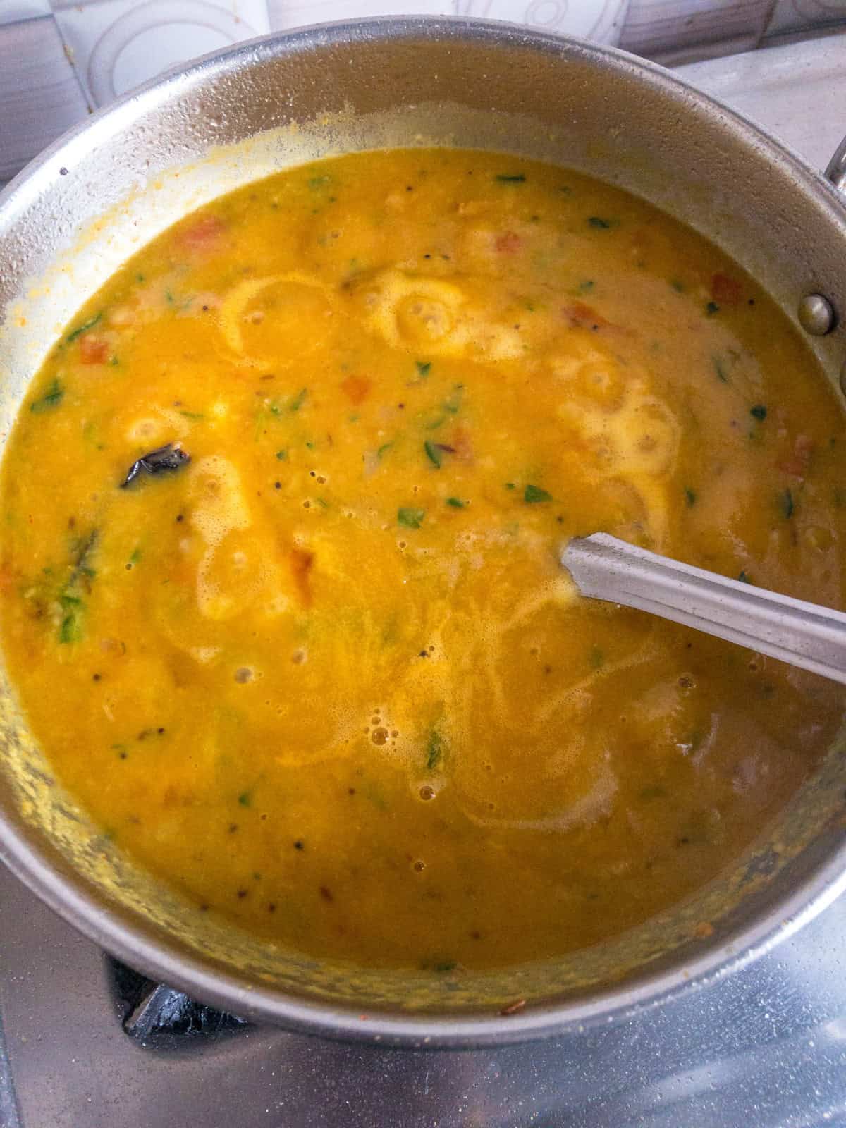 Finished methi toor dal simmering in a stainless steel pan with mustard seeds and tomato.