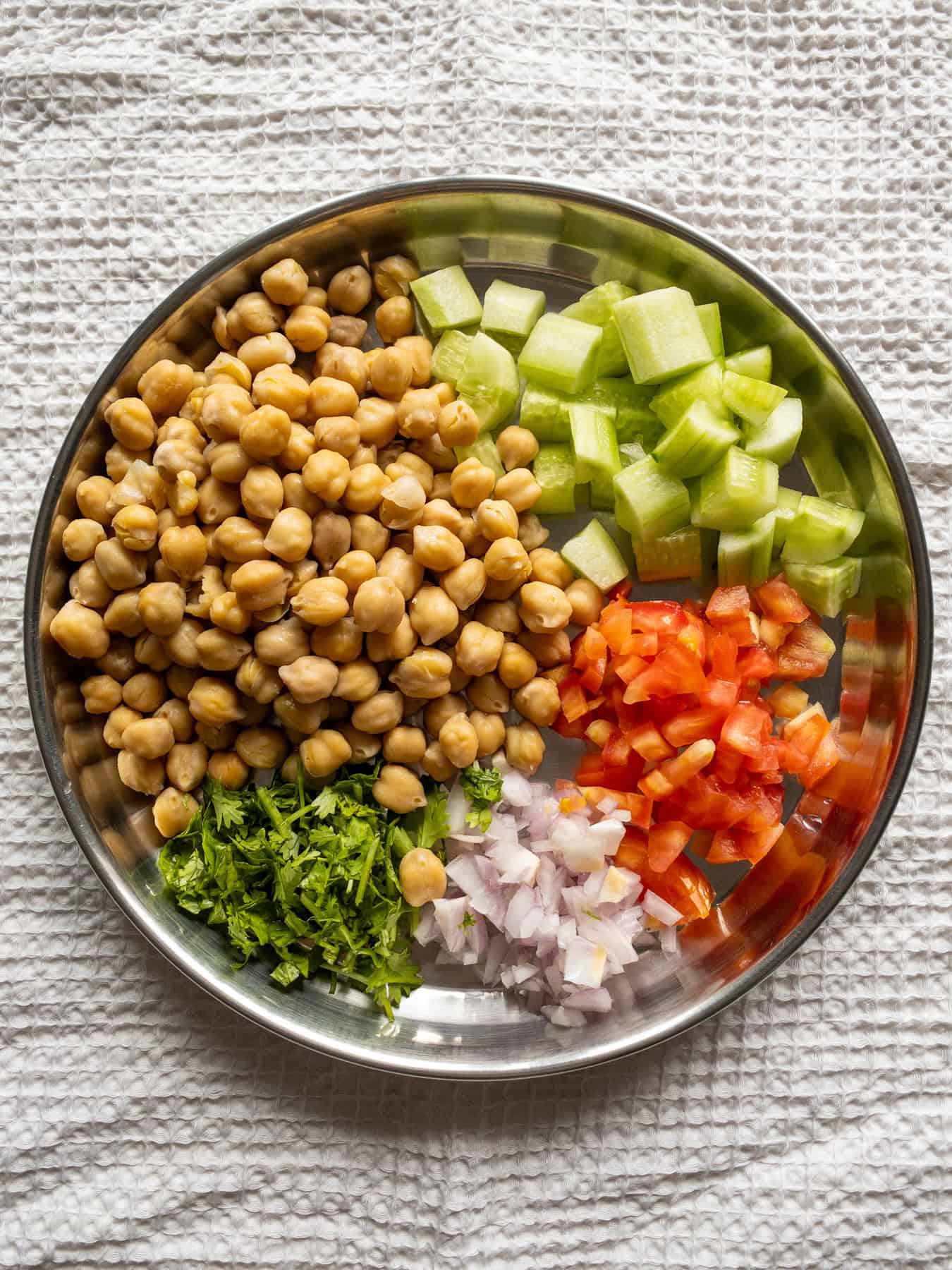 Cooked chickpeas with chopped cucumber, tomato, onion, and coriander arranged in a steel mixing plate.