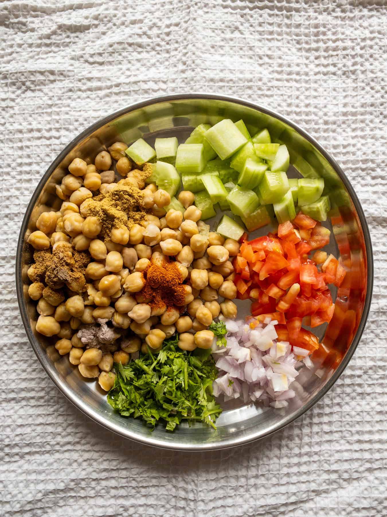 Cooked chickpeas and chopped vegetables topped with Indian spice powders in a steel mixing plate.