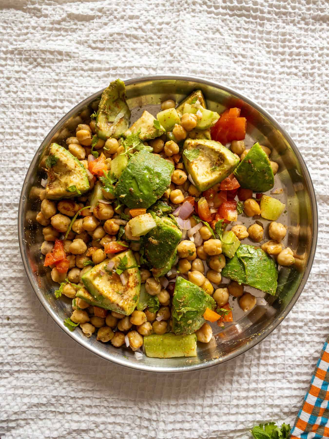 Chickpeas, avocado, cucumber, tomato, onion, coriander, and spices tossed together in a steel mixing plate.