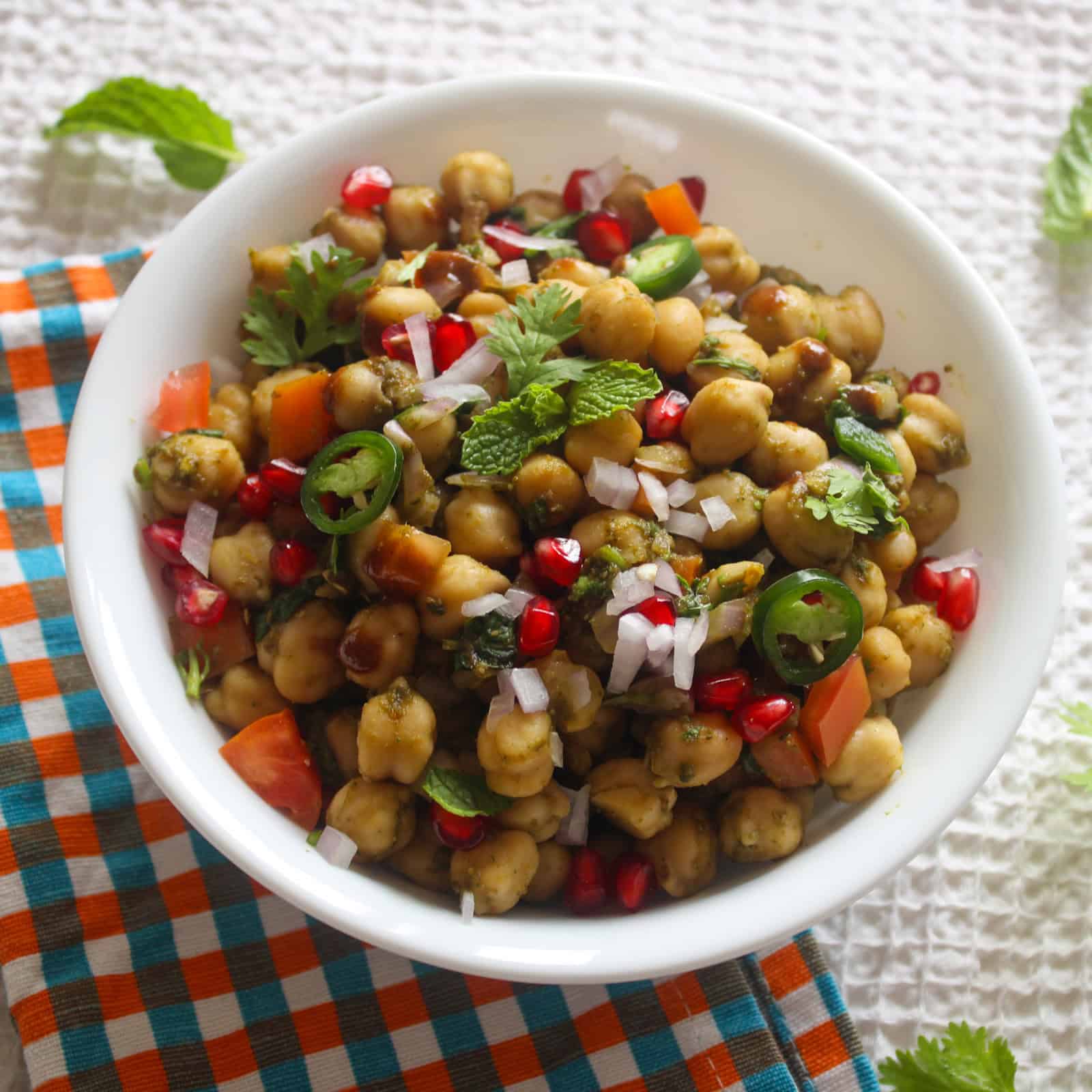 Close serving view of chana chaat with chickpeas, pomegranate, tomato, onion, fresh herbs, and sliced green chili.