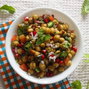 Chickpea chaat salad in a white bowl topped with pomegranate, chopped onion, cilantro, mint, tomato, and sliced green chili.