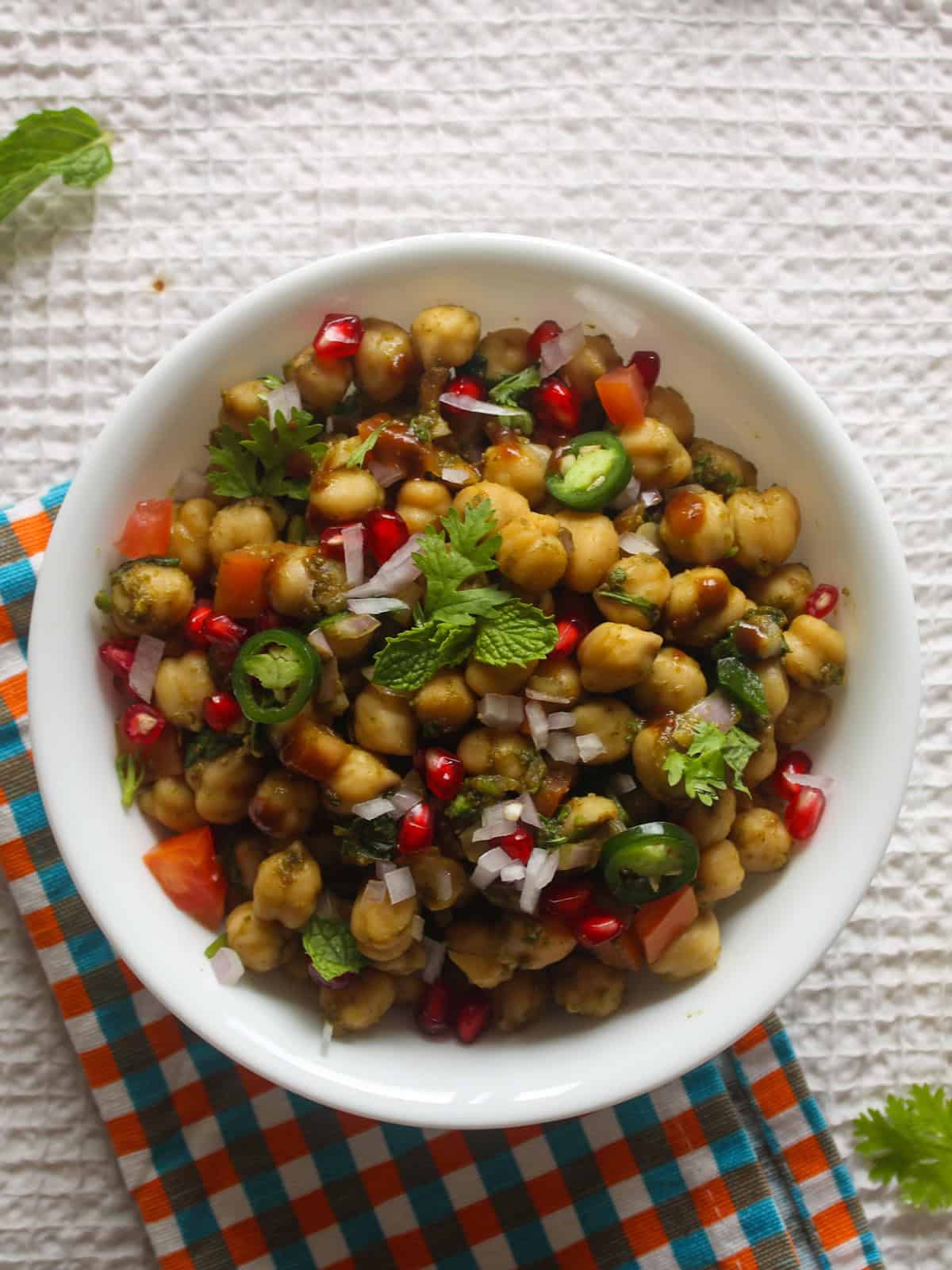 Chana chaat served in a white bowl with chickpeas, chopped vegetables, pomegranate, green chili, cilantro, and mint.