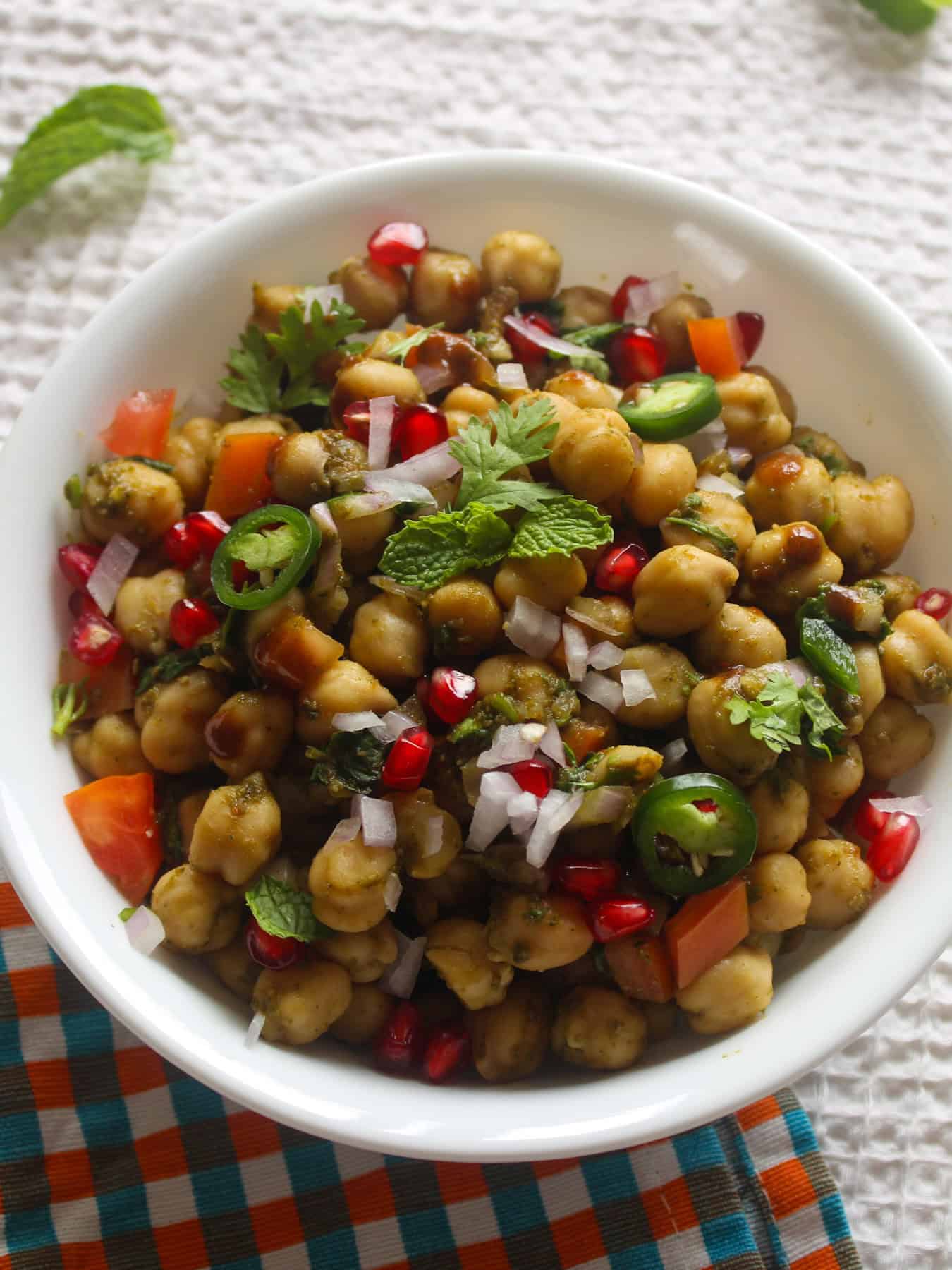 Close-up of Indian chickpea chaat topped with pomegranate, chopped onion, tomato, cilantro, mint, and sliced green chili.