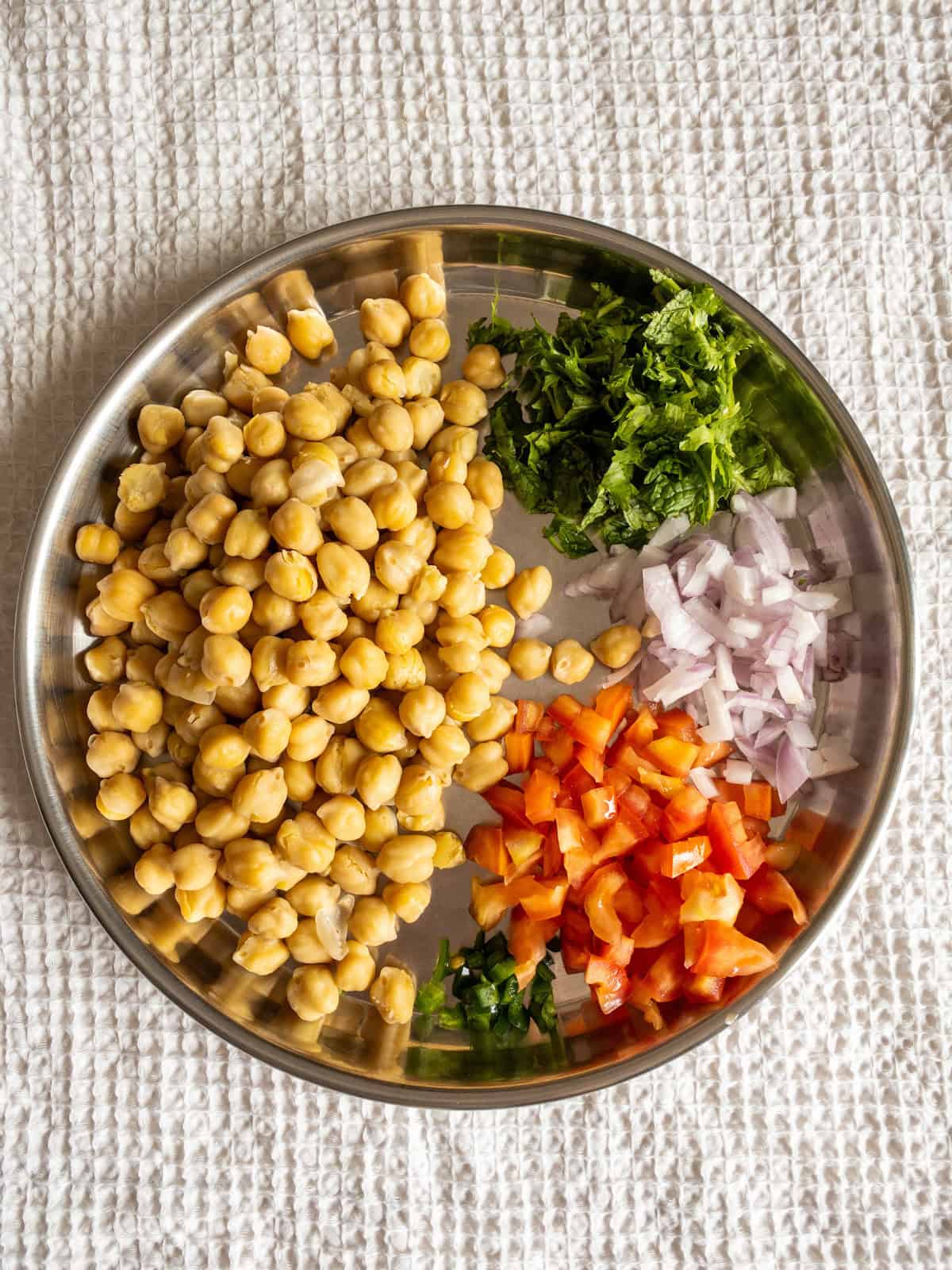 Cooked chickpeas with chopped tomato, onion, green chili, cilantro, and mint arranged separately on a steel plate.