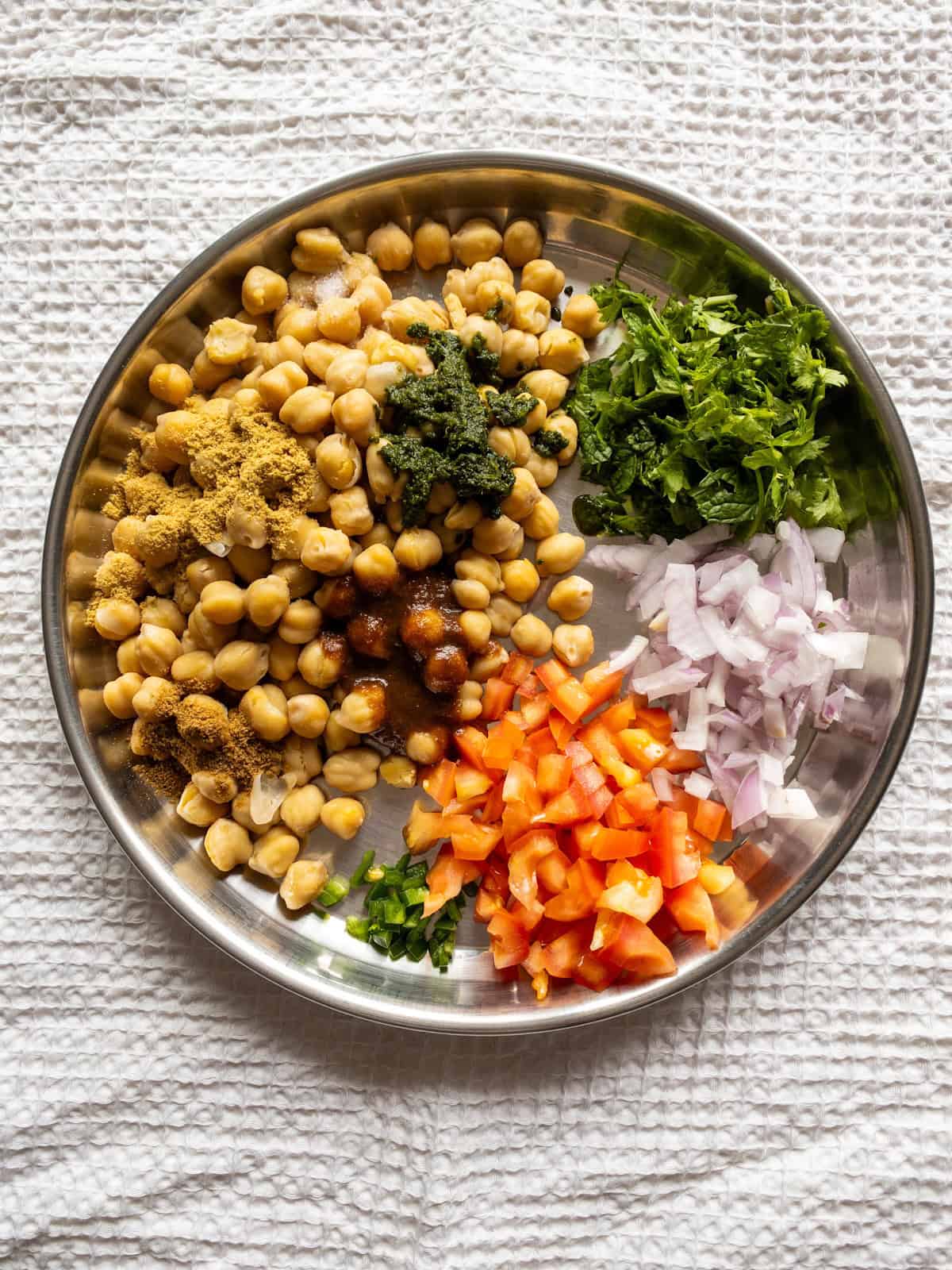 Cooked chickpeas with chopped vegetables, fresh herbs, chutneys, and ground spices arranged on a steel plate.
