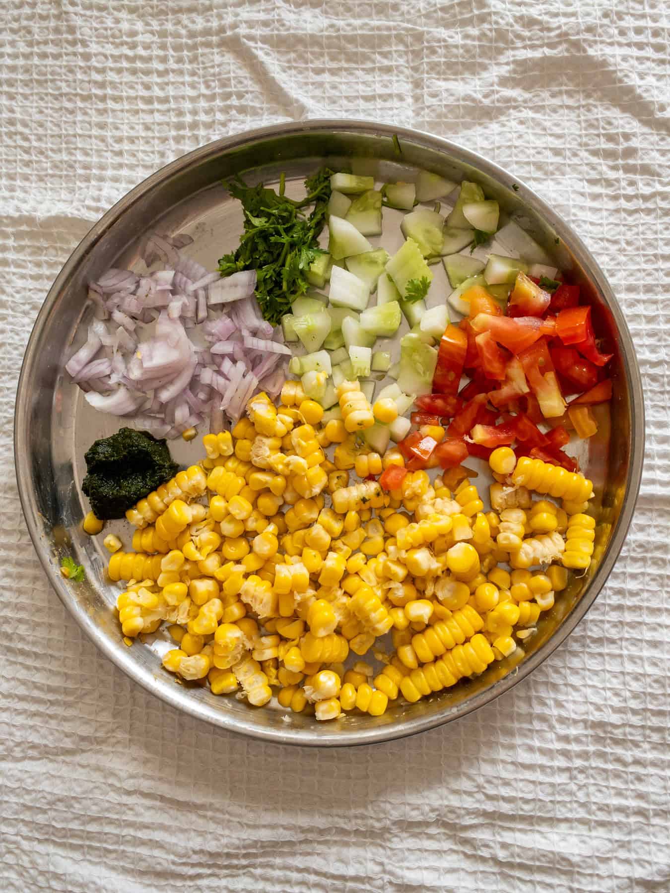 Steamed corn, chopped cucumber, tomato, onion, coriander, and green chutney arranged in a steel plate before mixing.