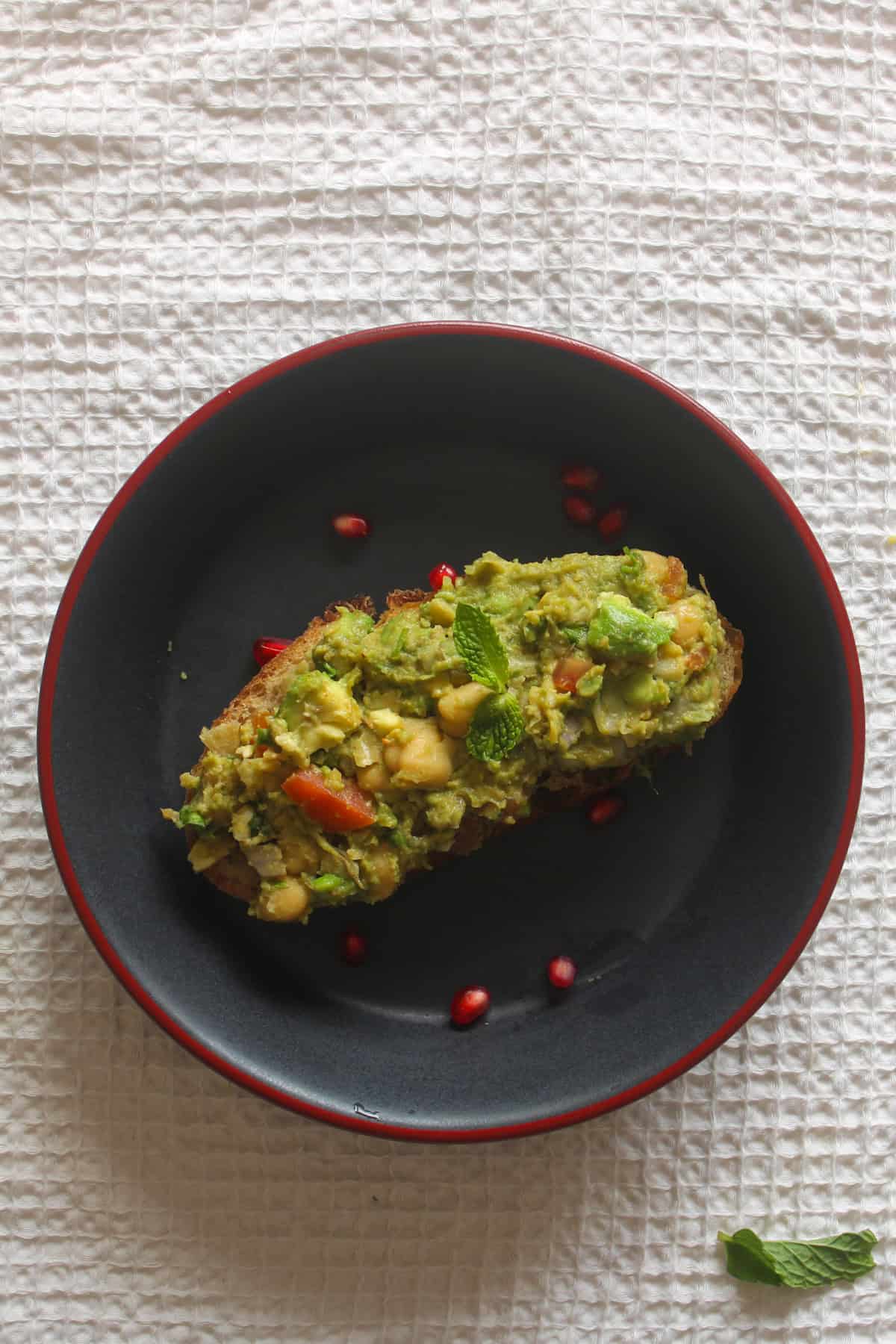 Indian spiced chickpea avocado toast served on a black plate with pomegranate arils and mint.
