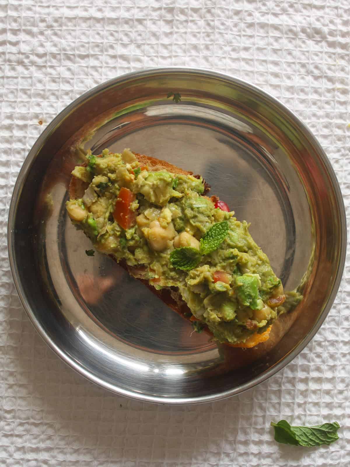 Sourdough toast topped with chunky chickpea avocado filling and mint on a stainless steel plate.