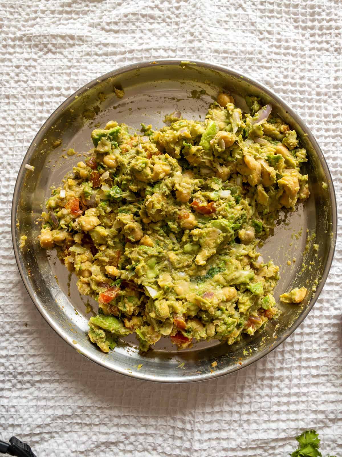 Chunky chickpea and avocado filling with onion, tomato, coriander, and spices in a stainless steel plate.