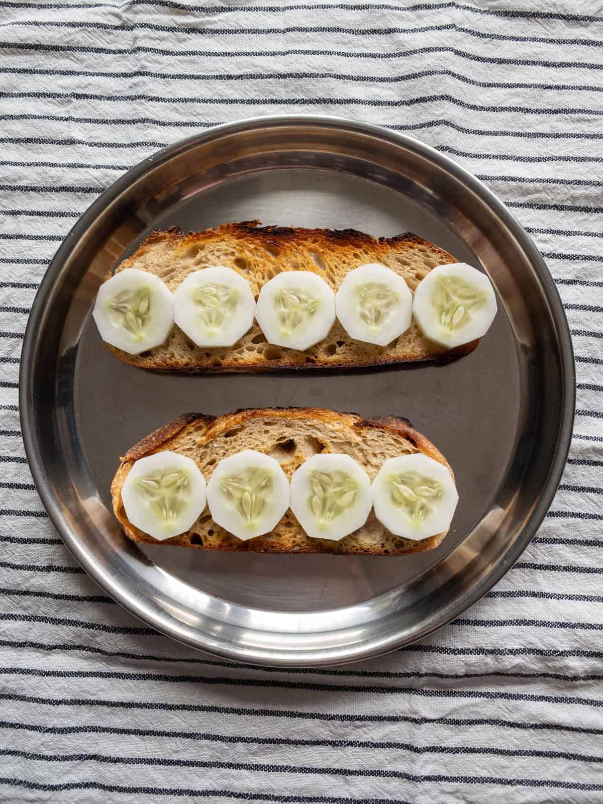 Two toasted sourdough slices topped evenly with round cucumber slices on a stainless steel plate.