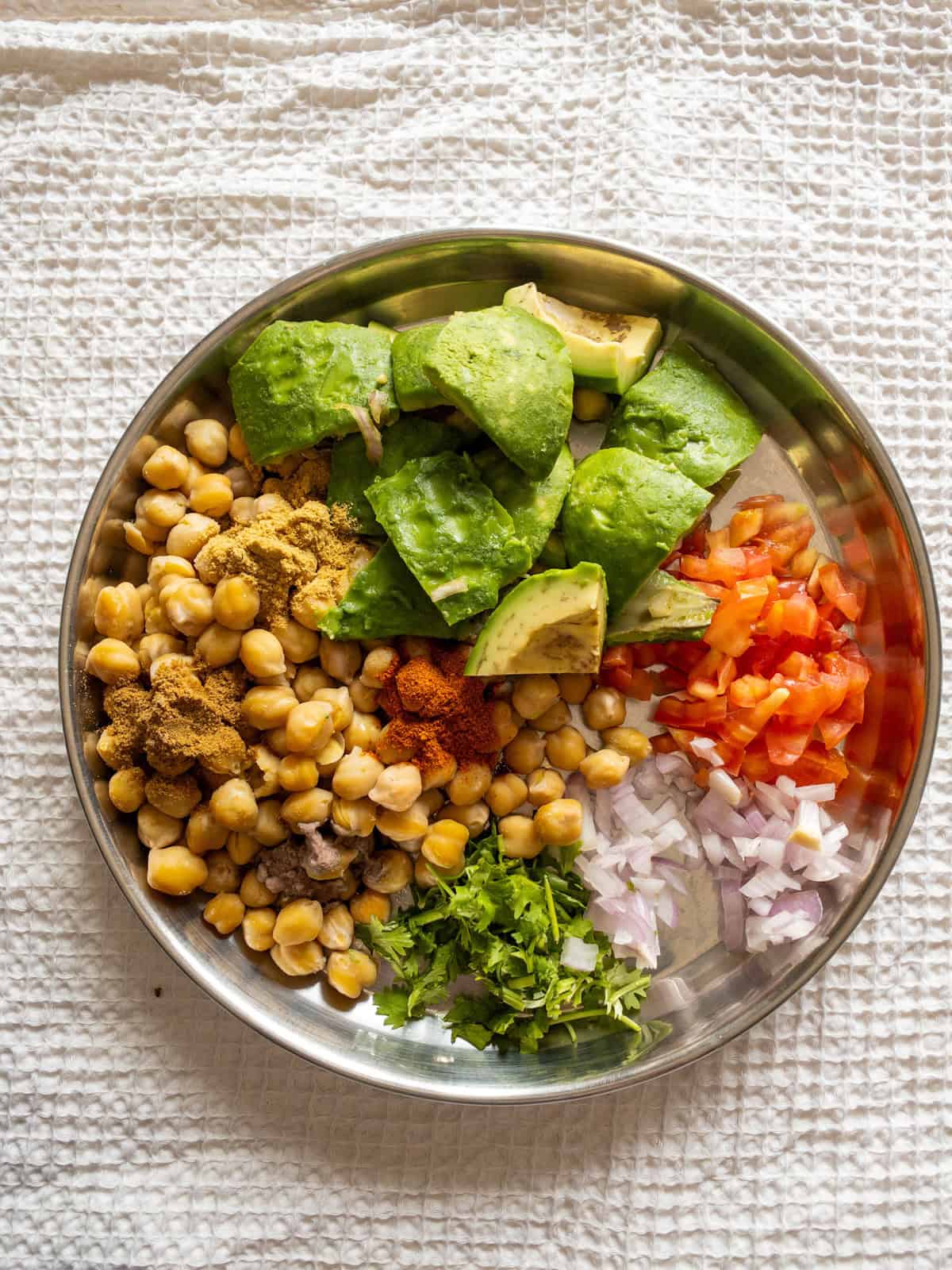 Cooked chickpeas, avocado, chopped tomato, onion, coriander leaves, and ground spices arranged in a stainless steel plate.