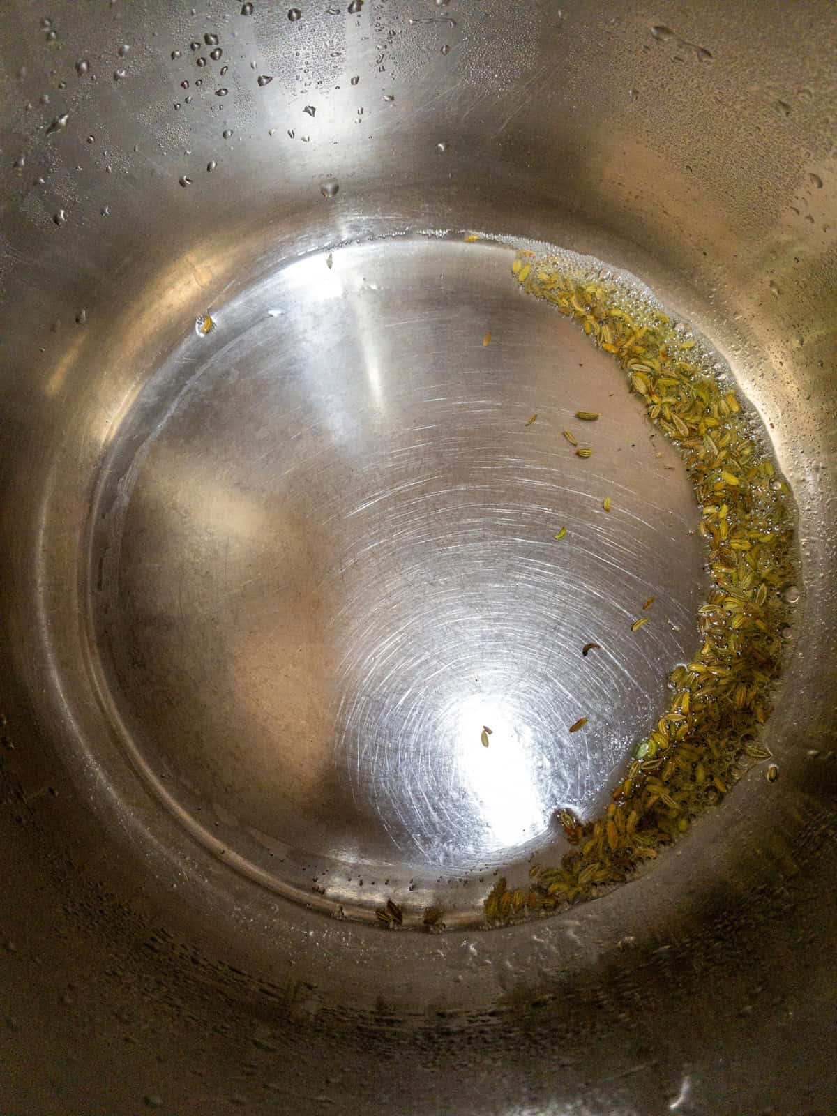 Fennel seeds warming in oil at the bottom of a stainless steel saucepan.